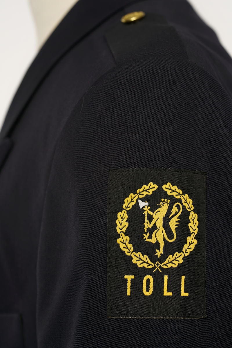 Mørkblå jakke enkelspent med tre riksløveknapper. Epauletter uten distinktsjoner. Tekstil marke på venstre arm, riksløve med hvit øks omgitt av eikeblader over TOLL. Hempe i nakke. Kypert stoff, blanding av polyester og ull. Jakke er foda med antatt syntetisk stoff, mørkblå.
Bukser av samme stoff.