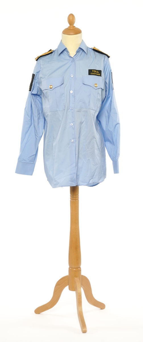 Lys blå langermet skjorte av et fintvevd stoff. Epauletter med tilsatt distinktsjon: krone over merkurstav over en kvist eikeblader (fire blader totalt), med gull bånd på begge sider. Tekstil merke på begge armer: krone over riksløve med hvit øks i rødt skjold omgitt av eikeblader over TOLL/CUSTOMS.