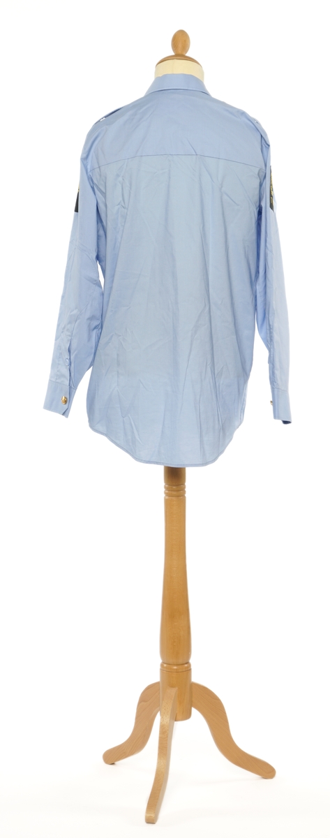 Lys blå langermet skjorte av et fintvevd stoff. Epauletter uten tilsatt distinktsjon. Tekstil merke på begge armer: riksløve med hvit øks omgitt av eikeblader over TOLL. Tekstil merke på venstre bryst, gul bokstaver på sort, 'TOLL'.