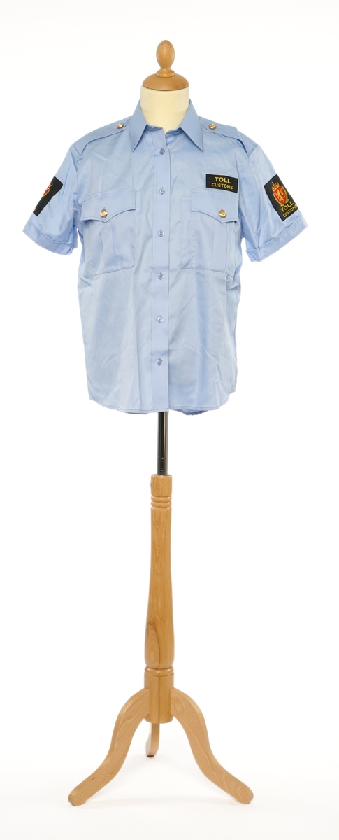 Lys blå  kortermet skjorte av et fintvevd kypert stoff. Epauletter uten tilsatt distinktsjon. Tekstil merke på begge armer: krone over riksløve med hvit øks på rødt skjold omgitt av eikeblader over TOLL/CUSTOMS. Tekstil merke på venstre bryst, gul bokstaver på sort, 'TOLL/CUSTOMS'.