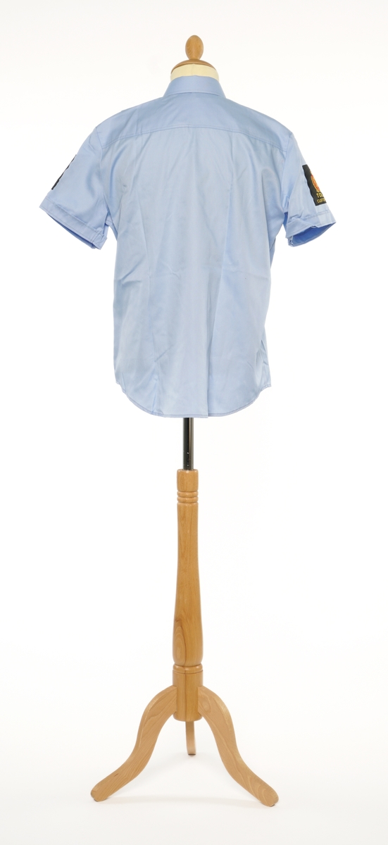 Lys blå  kortermet skjorte av et fintvevd kypert stoff. Epauletter uten tilsatt distinktsjon. Tekstil merke på begge armer: krone over riksløve med hvit øks på rødt skjold omgitt av eikeblader over TOLL/CUSTOMS. Tekstil merke på venstre bryst, gul bokstaver på sort, 'TOLL/CUSTOMS'.