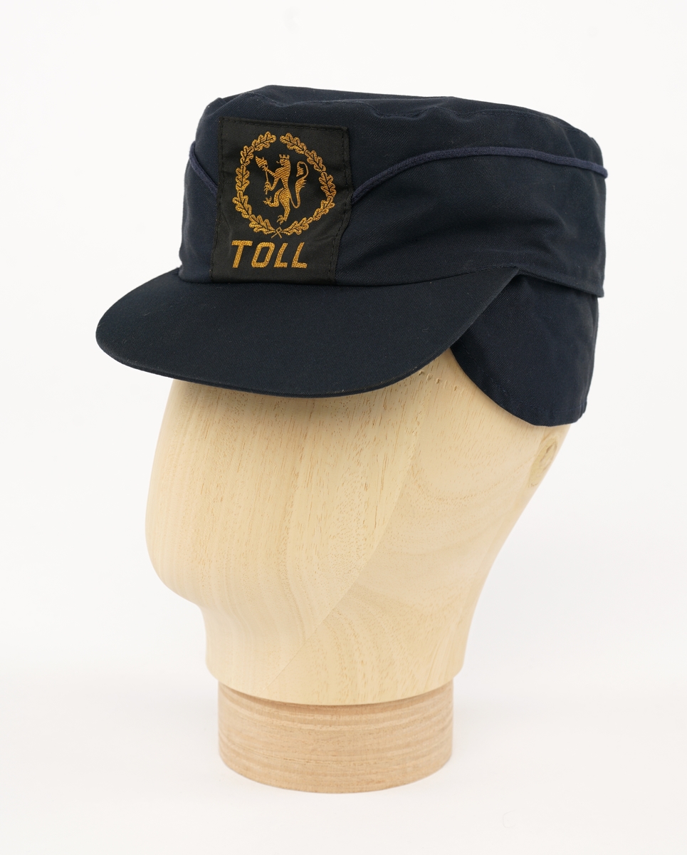 Mørk blå stoff hatt med visir. Tekstil merke på  forsiden, gul riksløve med øks omgitt av eikeblader på sort bakgrunn. Kypert stoff, antatt blanding av ull og polyester. Fora med lysere blå flanell stoff.