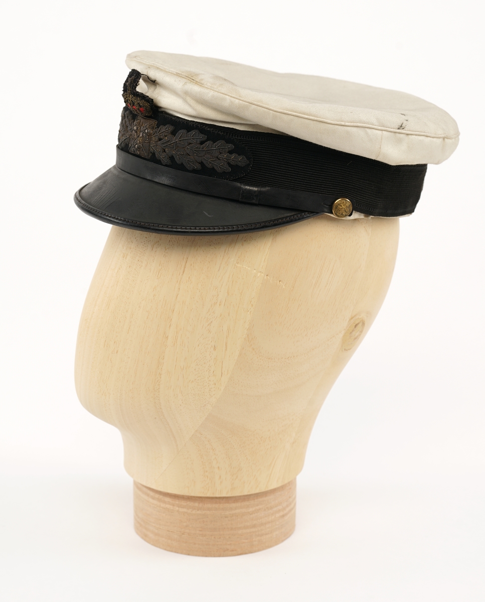 Hvit hatt med sort visir. Sort plast/lær strimmel/bånd over visir festet med riksløvesknapper på hver side. Merke på framsiden, krone over to eikekvister med tre stjerner i firkant.