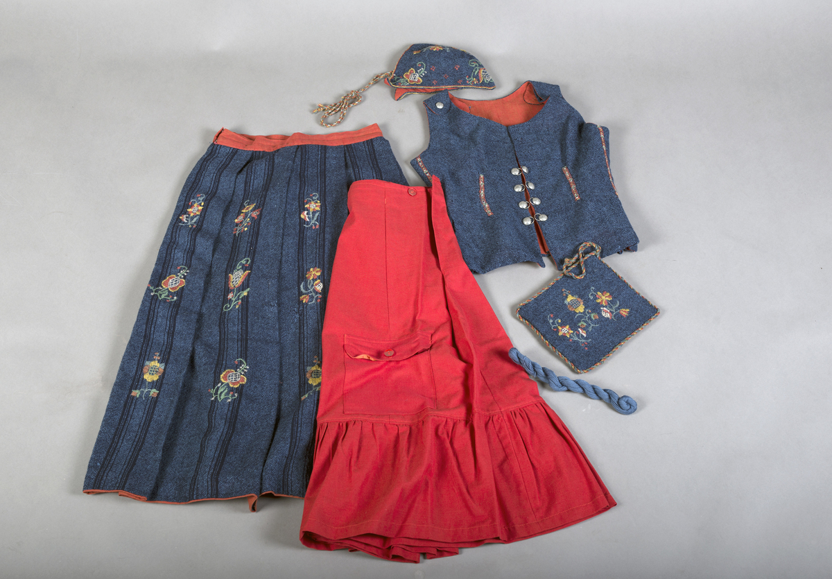 Rødt underskjørt med kappe og lomme, brodert linskjorte, brodert ull-skjørt, brodert ull-vest, brodert lue, brodert veske og blått brodérgarn i ull.