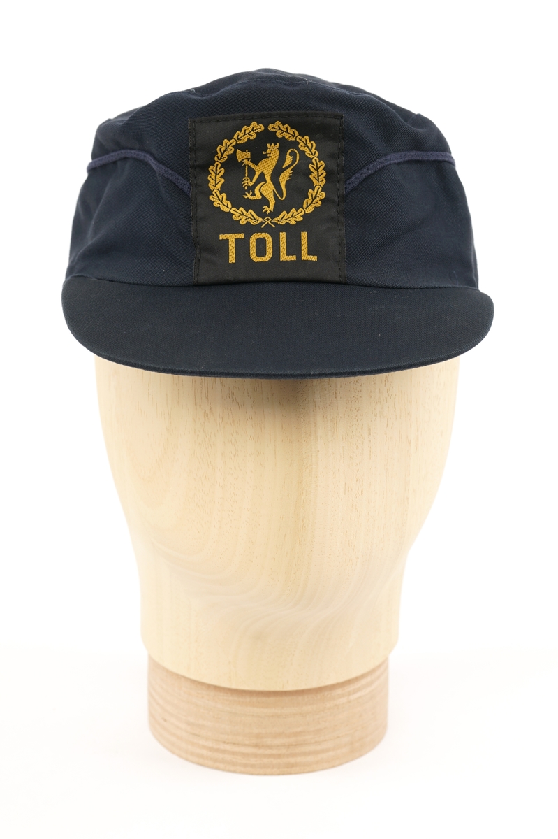 Mørkeblå  stoffhatt  med visir. Tekstil merke på forsiden, gul riksløve med øks omgitt av eikeblader på sort bakgrunn. Kypert stoff, antatt blanding av ull og polyester. Fora med lysere blå flanell stoff.