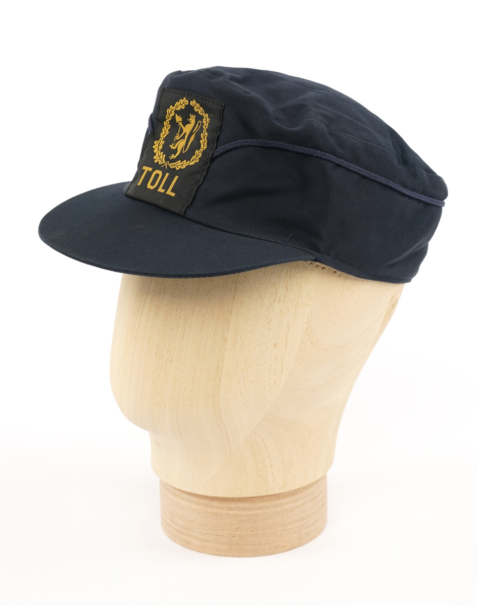 Mørkeblå  stoffhatt  med visir. Tekstil merke på forsiden, gul riksløve med øks omgitt av eikeblader på sort bakgrunn. Kypert stoff, antatt blanding av ull og polyester. Fora med lysere blå flanell stoff.