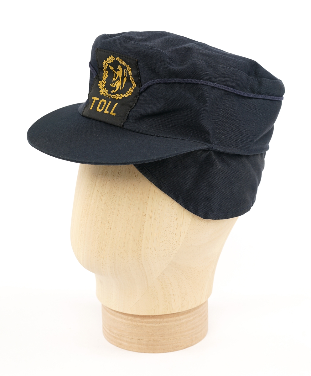 Mørkeblå  stoffhatt  med visir. Tekstil merke på forsiden, gul riksløve med øks omgitt av eikeblader på sort bakgrunn. Kypert stoff, antatt blanding av ull og polyester. Fora med lysere blå flanell stoff.