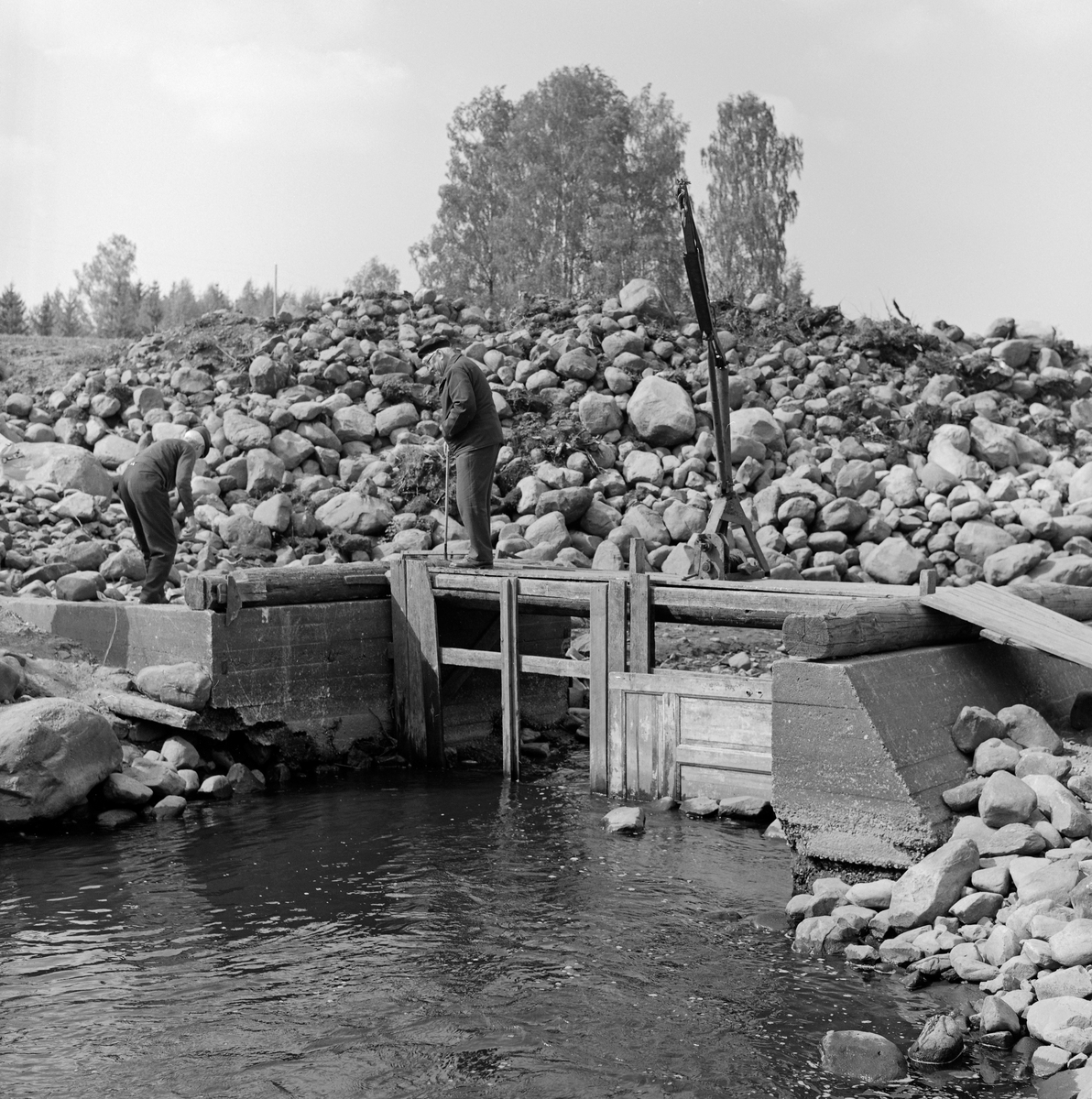 En «fiskebygning» ved Fossbakken ved Svartelva, som markerte kommunegrensa mellom Vang og Stange før vassdraget rant ut i Åkersvika og Mjøsa.  Fotografiet ble tatt i 1973.  Her hadde eierne av garden Nedre Kåterud i flere generasjoner hatt en hevdvunnen rett til å drive gytefiske – primært etter mort (Rutilus rutilus).  Vi ser at fiskebygningen var plassert i en del av elveløpet som var omgitt av støpte vanger, og der det var plass for to kuper (ruser), som ikke sto i elveløpet da dette fotografiet ble tatt.  På brua var det plassert ei kran.  På bildet ser vi (til venstre) Martinus Hovie.  Mannen som sto på brua er foreløpig ikke identifisert.  Da dette bildet ble tatt var fiskeplassen omgitt av mye stein, antakelig fordi hovedvegen mellom Oslo og Trondheim (E6) like ved var under utvidelse.