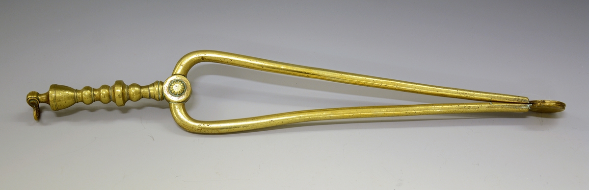 Ildtang av messing, del av peissett i tre deler. Bøyle øverst til oppheng, dreide kuler og blomst der armene deler seg. Avslutning på armene er to ovaler.