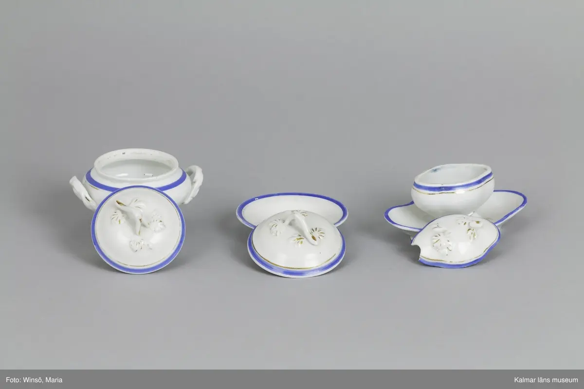 KLM 28568:5:1-8 Dockservis (matservis), fältspatporslin, vitt gods dekorerat med blå rand och guldkant (glansguld). Ostämplat. :5:1 Uppläggningsfat, oval form, guldkanten nött. :5:2 Uppläggningsfat oval form, guldkanten nött. :5:3 Tallrik, djup, guldkanten nött. :5:4-5 Tallrik, 2 st, flat tallrik, guldkanten nött. :5:6 Soppterrin på fot med lock, med våggrepe, svagt välvt lock med handtag i form av gren med blad, fals, guldkanten nött. :5:7 Karott med lock, locket svagt välvt med handtag i form av gren med blad. :5:8 Såsskål med lock, oval form, locket har urgröpning i ena sidan, avsett för slev, handtag har lossnat, guldkanten nött.
