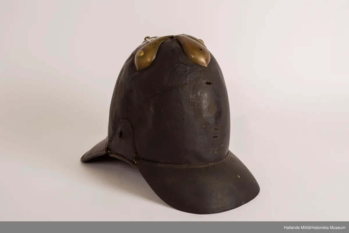 En s.k kask (hjälm) av svart styvt läder. Hjälmen är svagt sluttande upptill med förgylld plåt. Hjälmen har fram- och bakskärm. Förgylld plåt på baksidan som löper lodrätt från toppen av hjälmen ner på bakskärmen. Brunt läder på insidan.