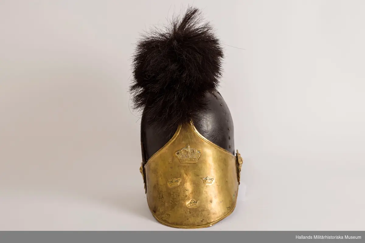 En s.k kask (hjälm) för manskap vid infanteriet. Hjälmen är gjord av styvt svart läder och har fram- och bakskärm. Framskärmens insida är mörkgrön. 
Svartlackade metalldetaljer på hjälmens ovansida samt tagelliggare med svart tagel. Förgylld framplåt med upphöjd relief i form av en stor sluten kungakrona samt tre mindre kronor. Förgyllda plåtdetaljer, reliefmönster i form av löv/blad, ovanför nackskärmen. Förgyllda fästen för hakrem i form av ett lejonhuvud på vardera sidan av hjälmen. Brunt läder på insidan och är märkt: 23 29½ med svart penna.