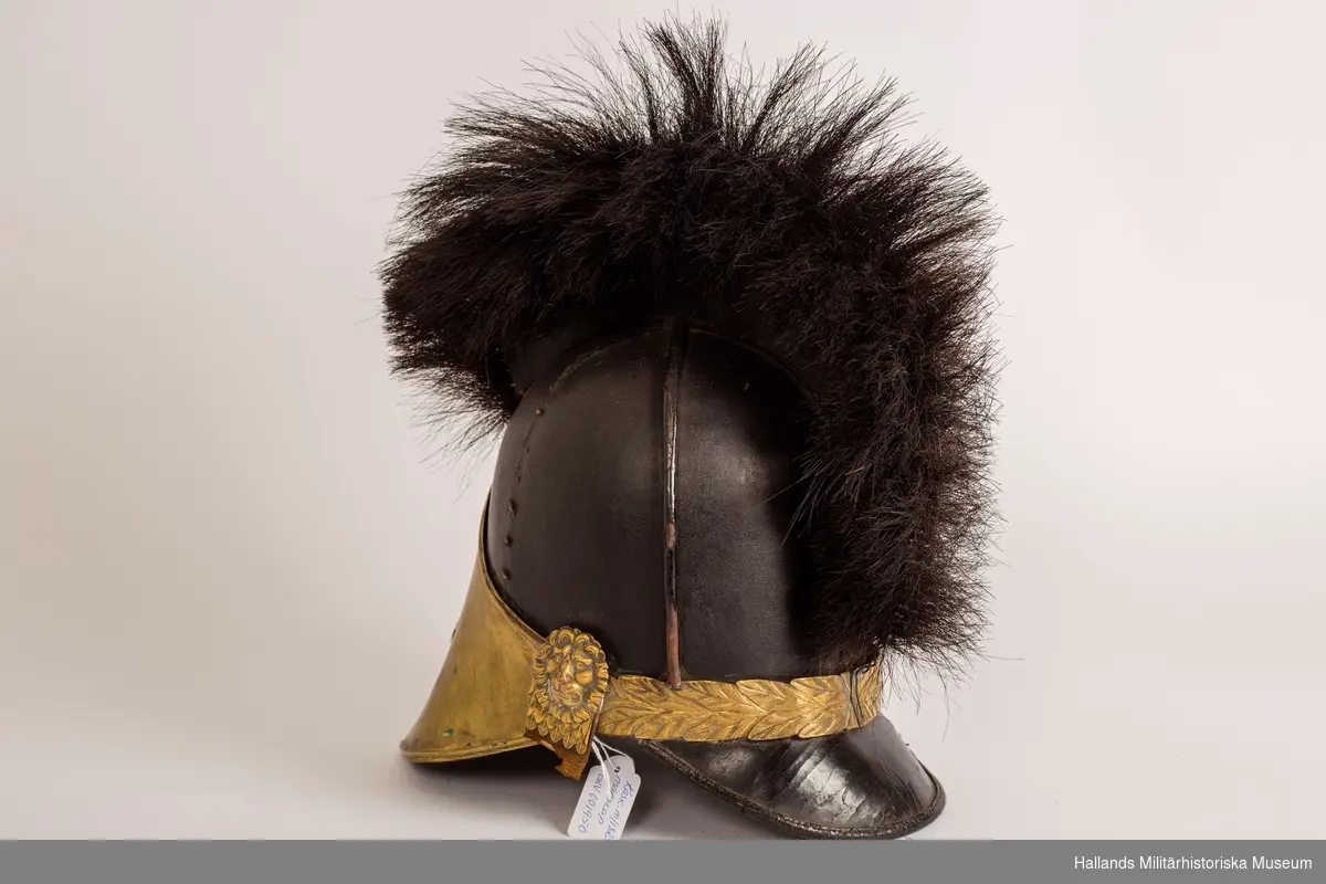 En s.k kask (hjälm) för manskap vid infanteriet. Hjälmen är gjord av styvt svart läder och har fram- och bakskärm. Framskärmens insida är mörkgrön. 
Svartlackade metalldetaljer på hjälmens ovansida samt tagelliggare med svart tagel. Förgylld framplåt med upphöjd relief i form av en stor sluten kungakrona samt tre mindre kronor. Förgyllda plåtdetaljer, reliefmönster i form av löv/blad, ovanför nackskärmen. Förgyllda fästen för hakrem i form av ett lejonhuvud på vardera sidan av hjälmen. Brunt läder på insidan och är märkt: 23 29½ med svart penna.