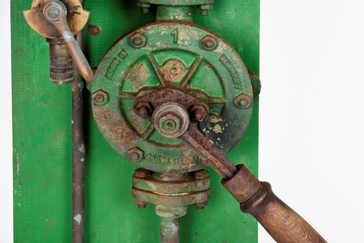 Oljepumpe med ventil montert på blå treplate. Tanken tar 5 liter. Feste for å sette en kanne e.l. under tanken. 

Ventil er produsert i UK, merke Runwell No 0
Oljepumpe fra Norsk Brendselsolje. Dette ble senere Noroil og deretter Statoil. Se: https://no.wikipedia.org/wiki/Norol