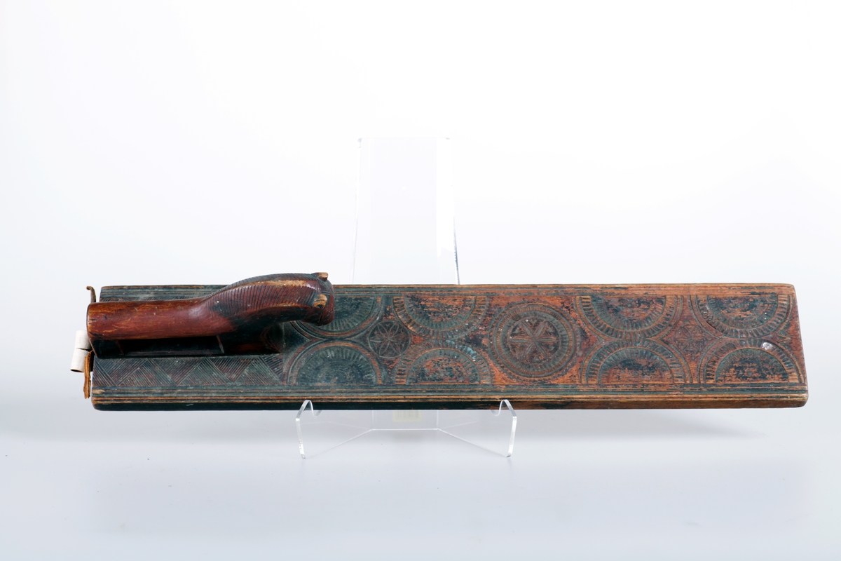 Mangletreet er malt rødt og sort. Karveskurd. Hestehåndtak, smal profil på sidene. Sikk sakk ornamentikk ved hestehåndtaket. Dekorert med sirkler og halvsirkler, og seksbladsroser i sirklene. Lærhempe i enden. Håndtaket festet med jernskruer.