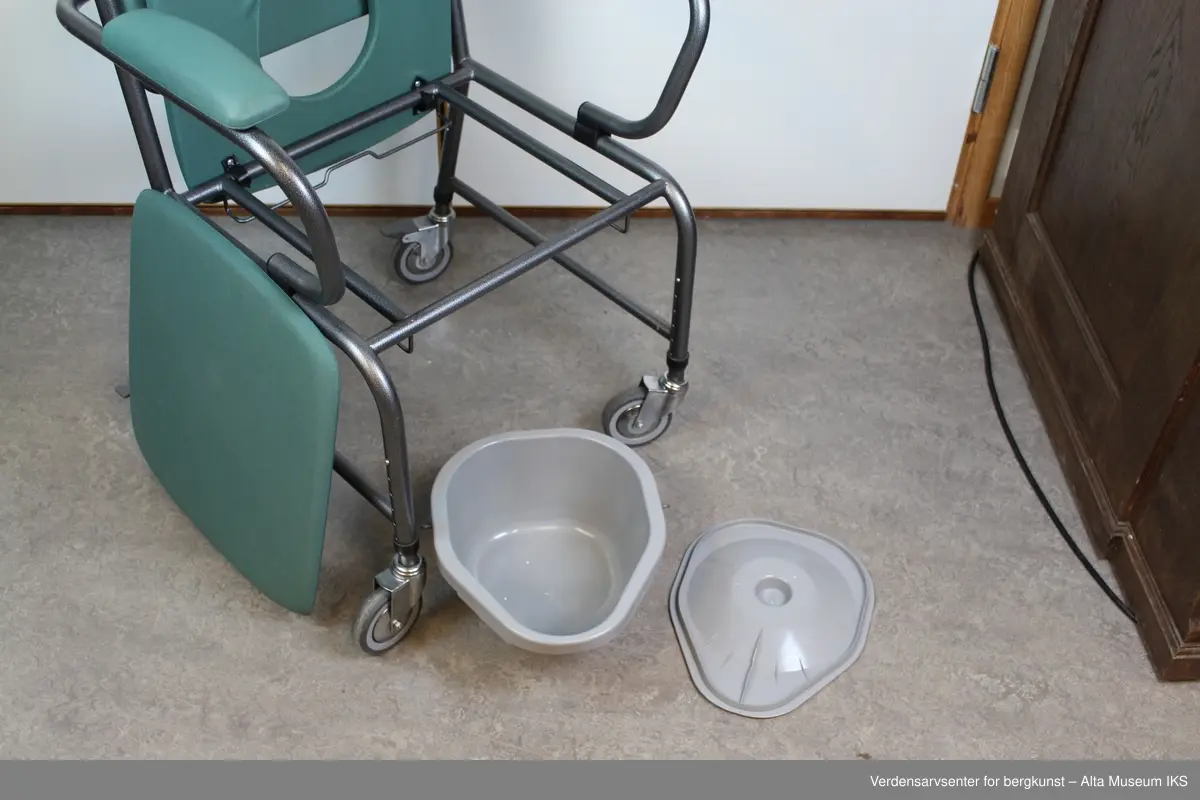 Plastbelagt stålrørstol på hjul. Med avtakbart sete over et dosete og dobøtte. Setet, ryggen og armlenene har puter med et grønnt trekk. Har håndtak for å trille stolen.