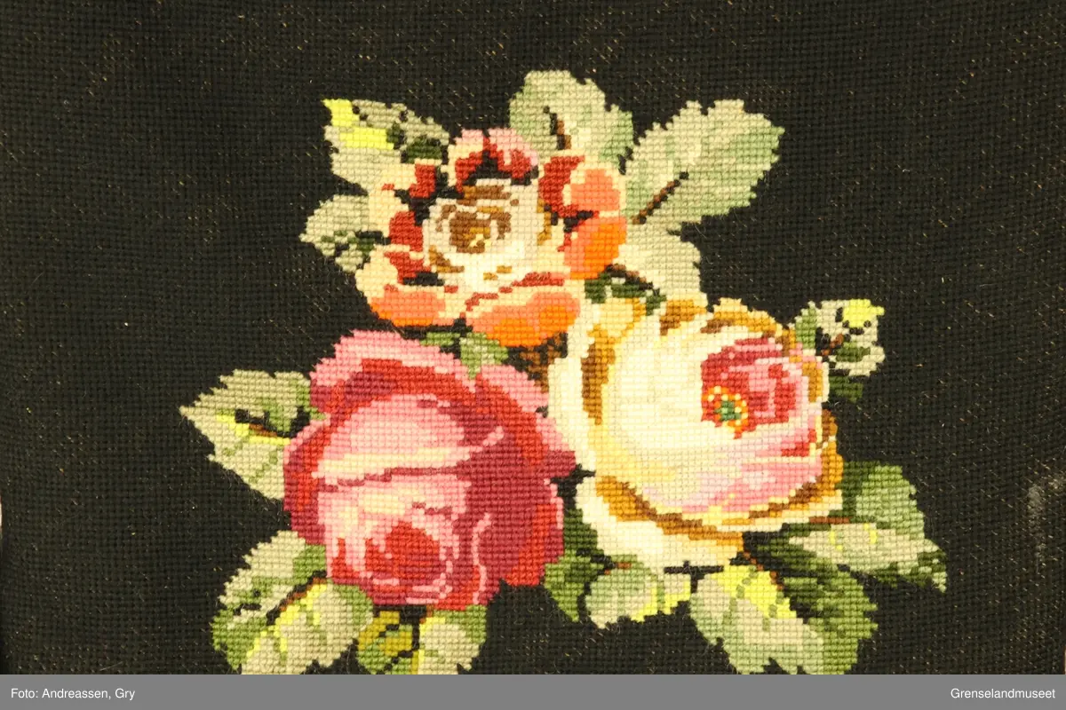 Nakkeputen og ryggputen er i svart aida med brodert rosemønster. Gyngestolen har gulltegninger på setet og rygg.