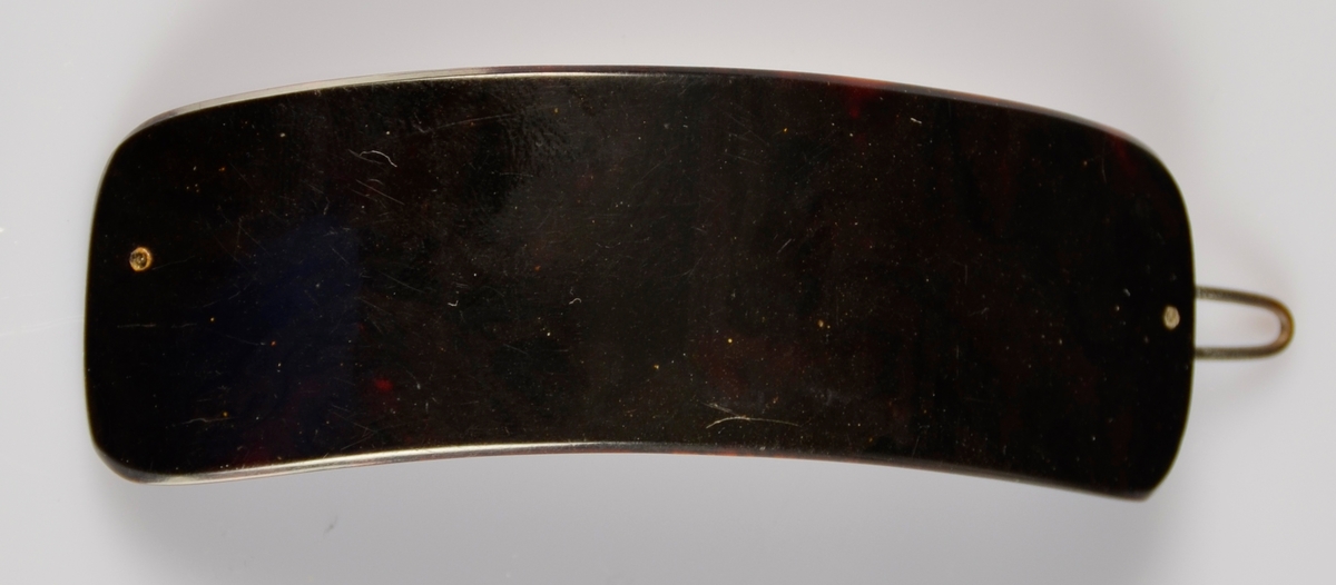 Hårspenne med noko boga form, med metall klype på baksida. Mørk brunt plastliknande materiale.