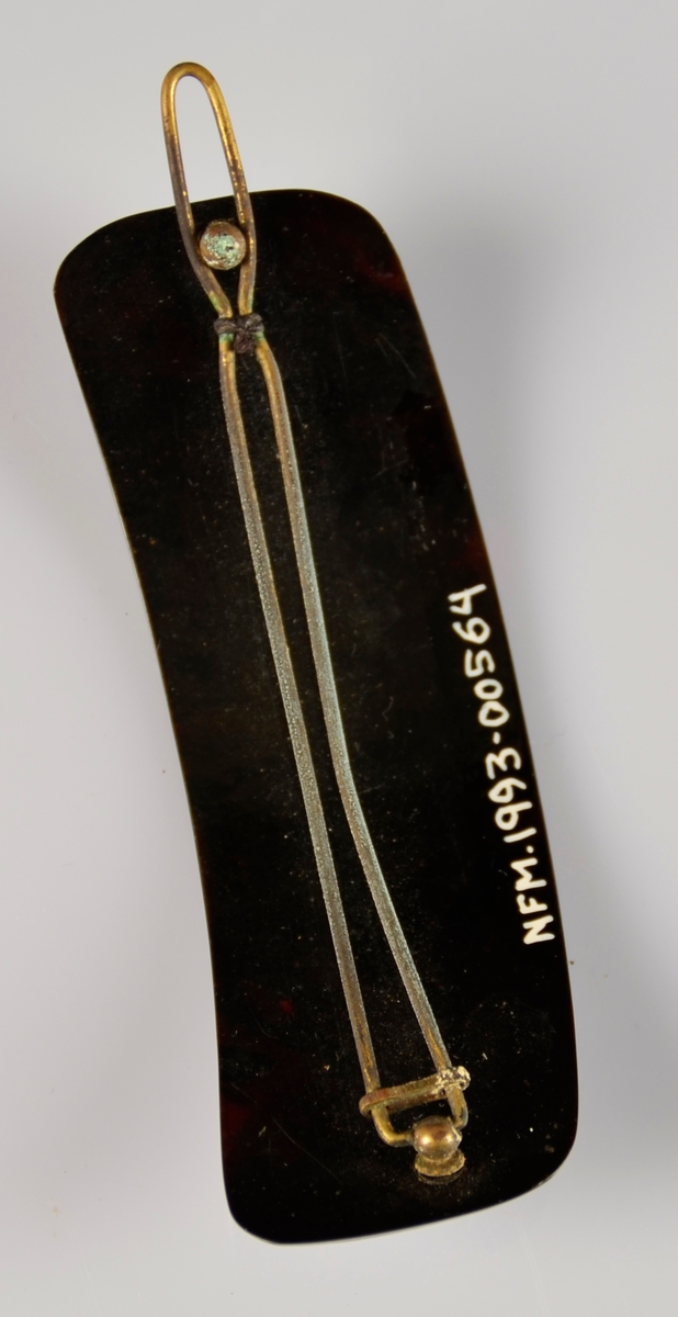 Hårspenne med noko boga form, med metall klype på baksida. Mørk brunt plastliknande materiale.