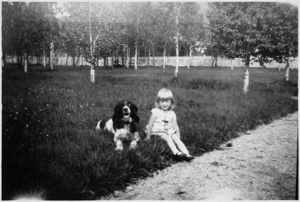 Jente og hund sitter på gresset i Byparken, Mosjøen. Kan være nasjonaldagen i 1934 (bildeserie).