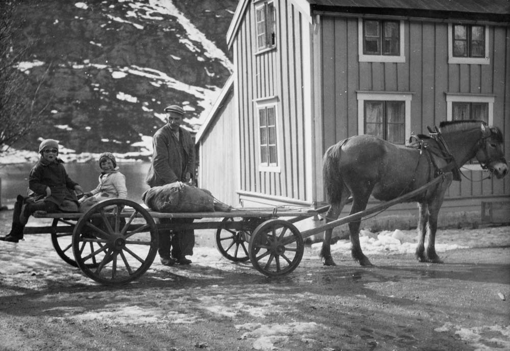 Hest og vogn med to barn på vognen. En mann står og holder oppsyn. Ved Fru Haugans Hotell.