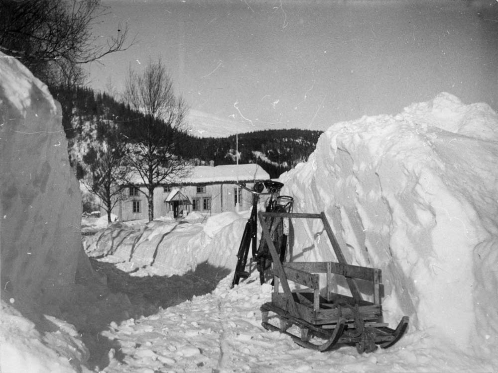 Hvitt hus med flaggstang under ås. Sykkel og kjelke ved snøskavl.