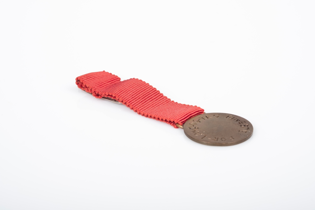 En rund medalje i metall med rødt bånd. På medaljens bånd er det påfestet en sikkerhetsnål. Medaljen har H7-symbolet. På baksiden av medaljen er det tekst som ser ut til å være inngravert på hånd.