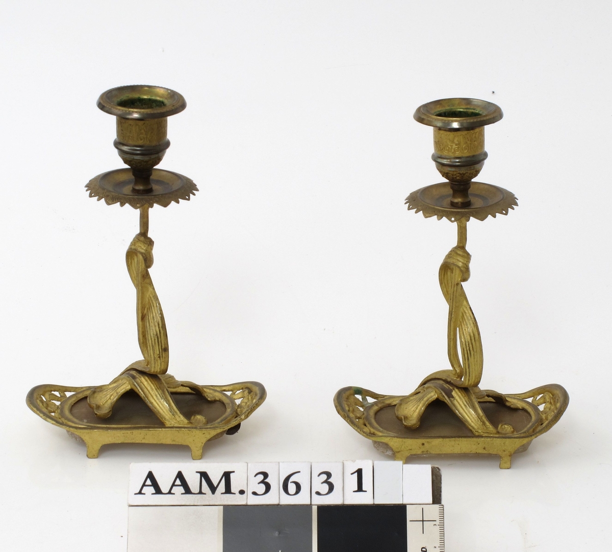 To like lysestaker av messing og polert stein. Avlang fot på fire ben med rette  langsider og avrundede, oppbøyede  kortsider. Skaftet vokser som to sammenflettede blad opp fra en  bladklynge på foten. Holderog dryppeskål med profileringer og bladornamenter.