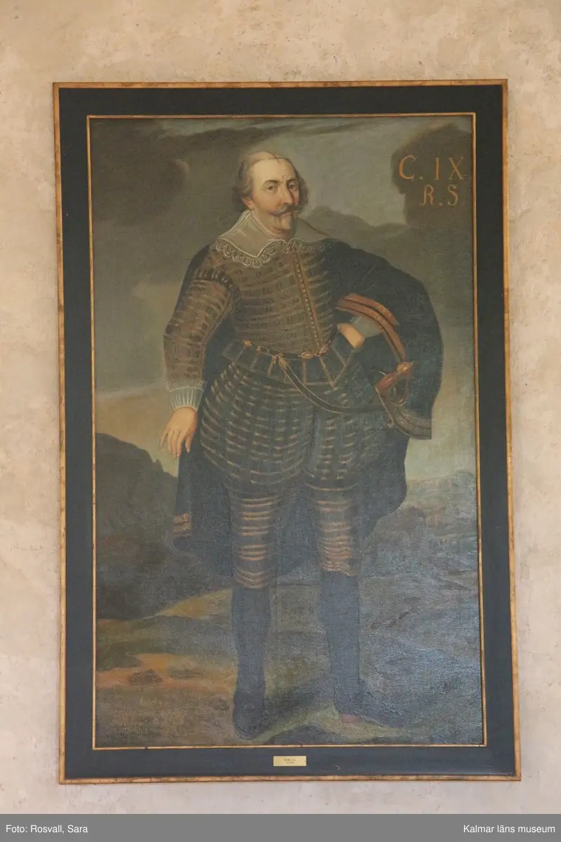 Karl IX i helfigur, stående, i mörk dräkt med guldstickningar, mantel med guldbroderier, spetskrage samt spetsar vid handlederna. Värja, svarta strumpor och skor med spännen.