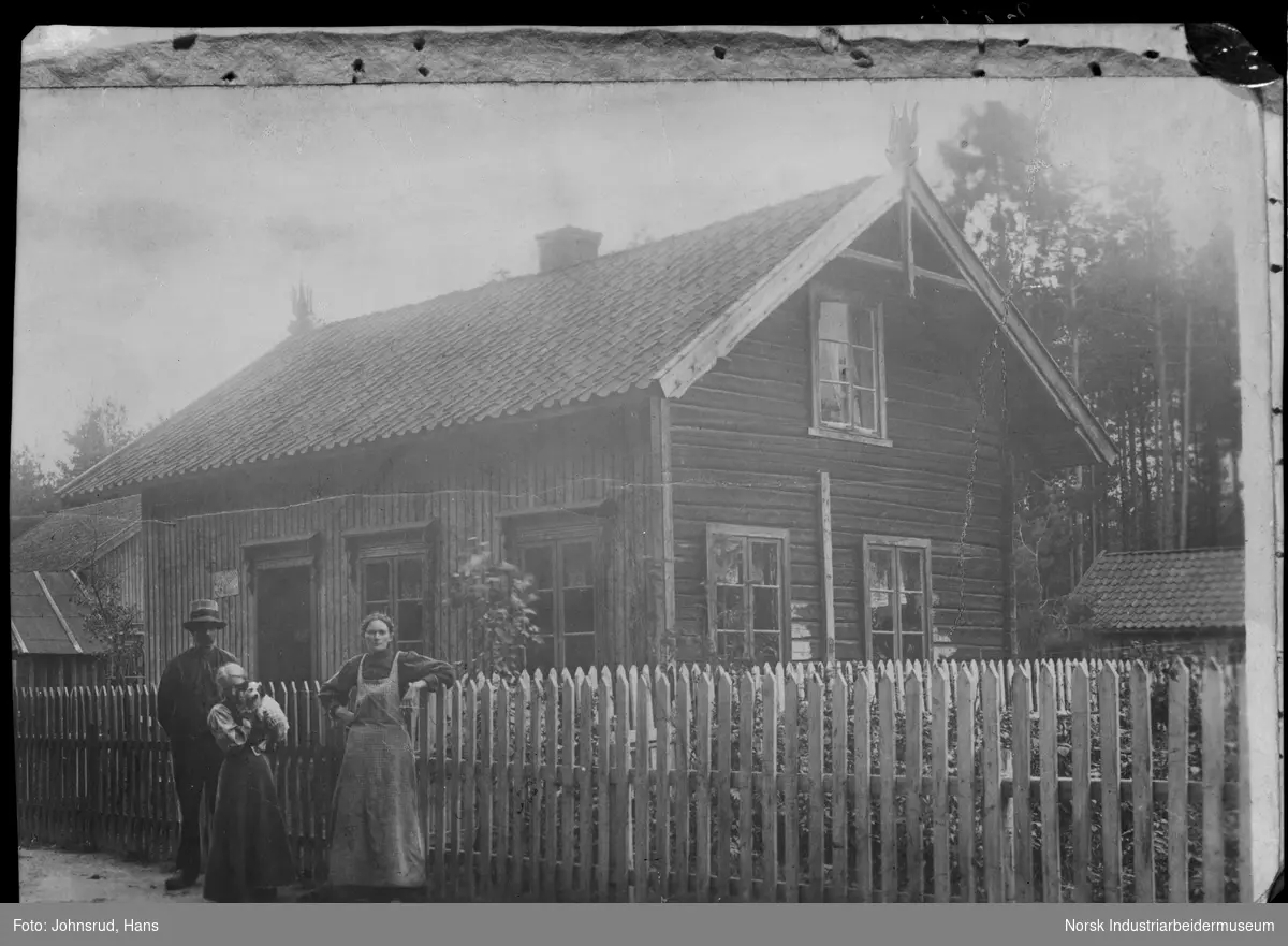 Avfotografert eldre foto. Kvinne, mann og jente stående utenfor gjerde til bolighus. Jenta har en hund i armene.