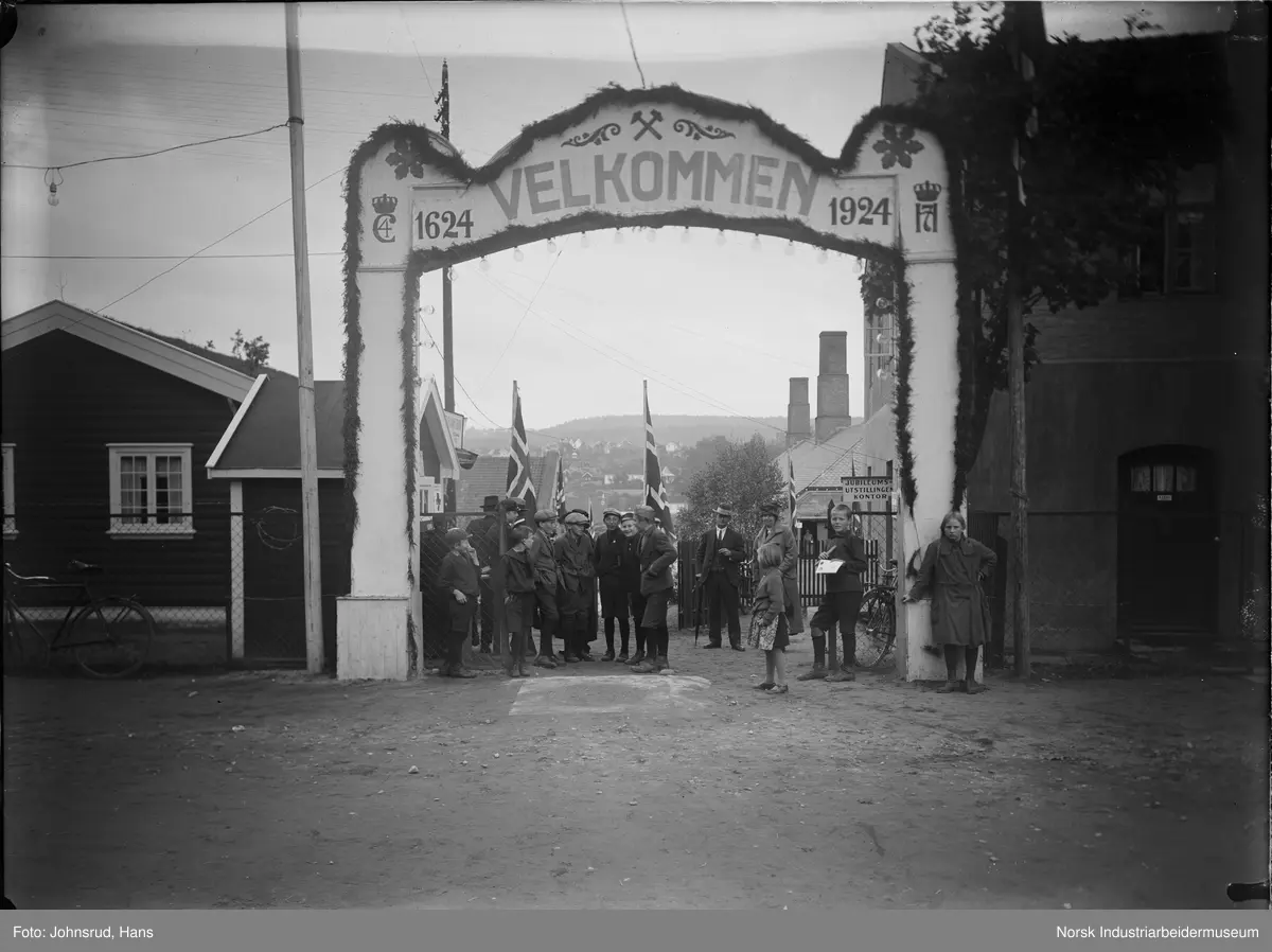 Fylkesutstillingen Kongsberg 1924. Portal med teksten "1624 Velkommen 1924". Barn og voksne stående under portalen, norske flagg på sidene.