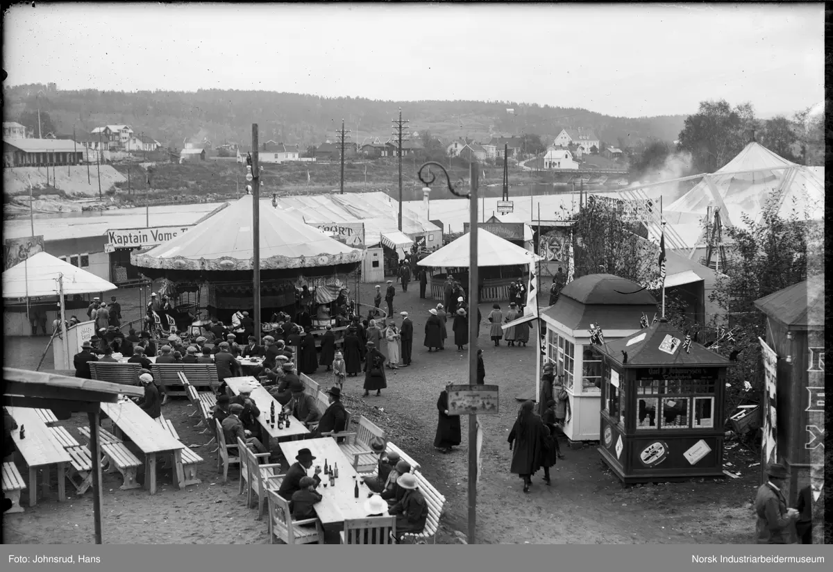 Fylkesutstillingen Kongsberg 1924. Mennesker ser på utendørs utstillinger og aktiviteter. Telt satt opp på plass ved elva Laagen. Mennesker sittende ved bord med flasker på bordet.