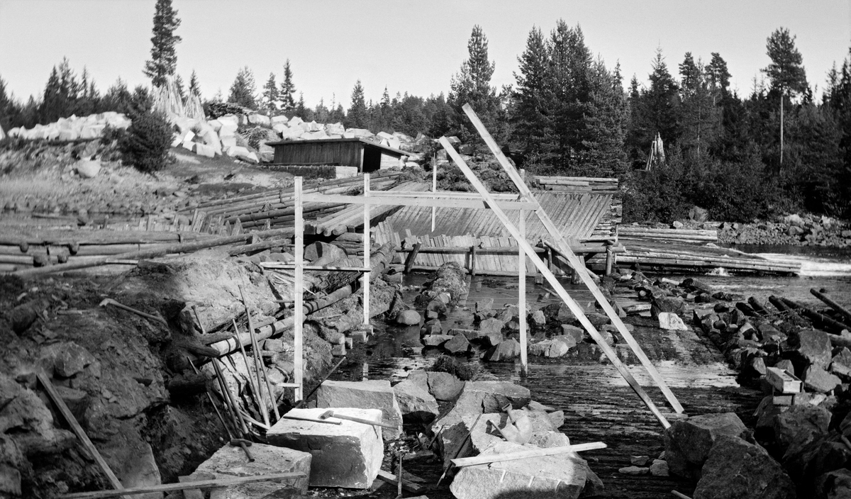 Første fase i ombygginga av Kjellåsdammen i Flisavassdraget i Åsnes i Solør (Hedmark) i 1915.  Fotografiet er tatt seinhøstes, på et tidspunkt da vannføringa i Flisa var meget lav, slik at konstruksjonene var lett tilgjengelige for anleggsarbeiderne.  Da dette bildet ble tatt var de «gamle» konstruksjonene - en steinfylt tømmerkistedam - under demontering.  På elvebrinken på sørsida av vassdraget ser vi en god del steinblokker som var kjørt fram til vassdraget med sikte på at de eksisterende steinfylte trekonstruksjonene skulle erstattes av solidere og mer bestandige konstruksjoner i gråsteinsmur.  

Kjellåsdammen var en såkalt atthaldsdam som kunne demme opp den nedre delen av Flisavassdraget i en lengde på opptil fem kilometer.  Den dammen det avbildete anlegget avløste var bemannet med to damvoktere så lenge det pågikk fløting i Flisa.  Tidlig på 1900-tallet var fløtingskyndige i tvil om hvorvidt det svarte seg å vedlikeholde og bemanne en føtingsdam på dette stedet.  Det ble snakket om at Kjellåsdammen samlet lite vann, og at det av den grunn kunne være aktuelt å rive den så snart den nedenforliggende delen av vassdraget var bedre «opprenset».  I stedet ble det altså til at man etter rivinga av den gamle dammen bestemte seg for å bygge en ny dam, denne gangen en murkonstruksjon av digre natursteinsblokker.  Den nye dammen ble bygd i perioden 1916-24 (jfr. bl. a. SJF.1989-02336).

Den første dammen ved Kjellåsfossen ble oppført på midten av 1800-tallet - jfr. fanen «Opplysninger».