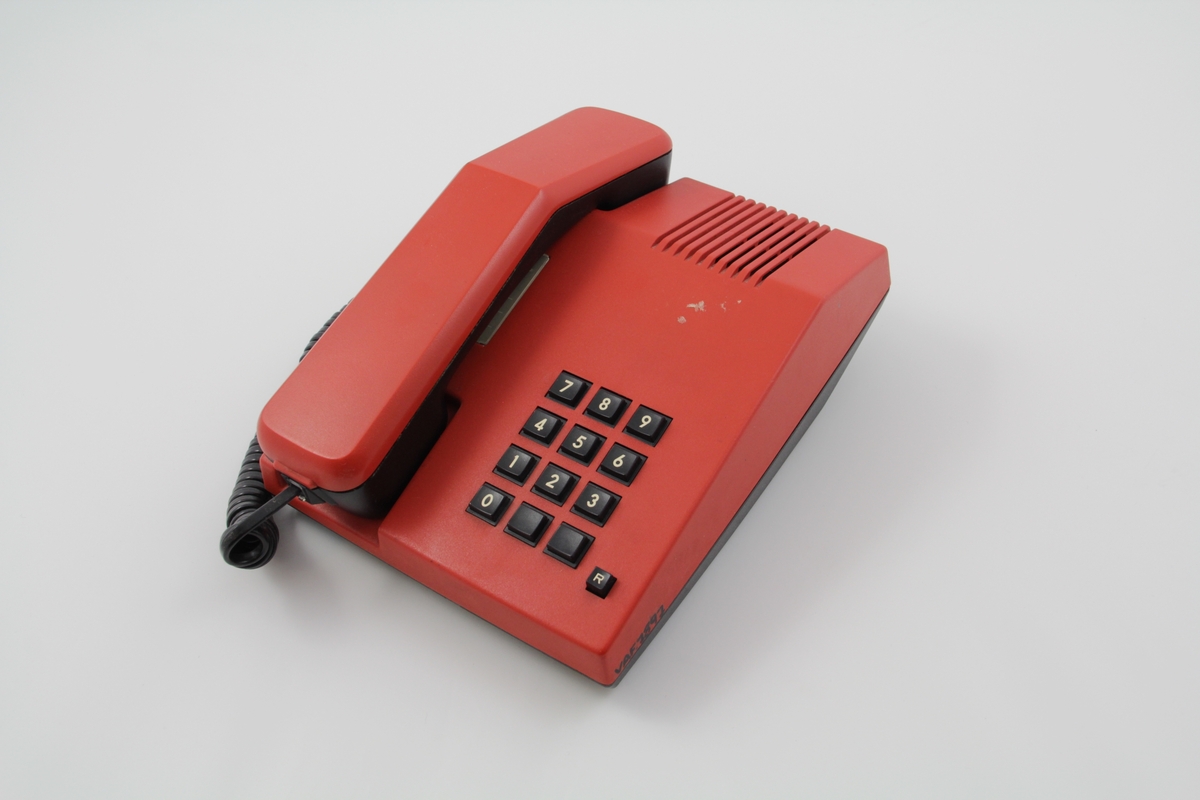 Rød telefon med tastatur. Telefonen er rektangulært formet med skråstilt front med tastatur, og har en fordypning for plassering av tale-/hørerør. Ledningsdel med egen telekontakt. På undersiden to reguleringsknapper: "tone". På overdeksel høyttaler/lyd-forsterker "vilum"