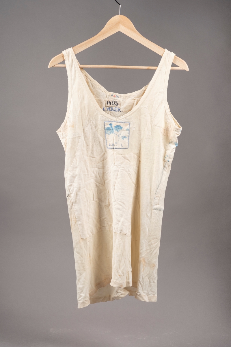 Hvit ullsinglet stoppet og lappet etter skader. Det er brodert inn initialer og navn på ulltrøyen i blå tråd. Noen av lappene har blomstermønstret stoff. Fangenummer er skrevet på i nakken.