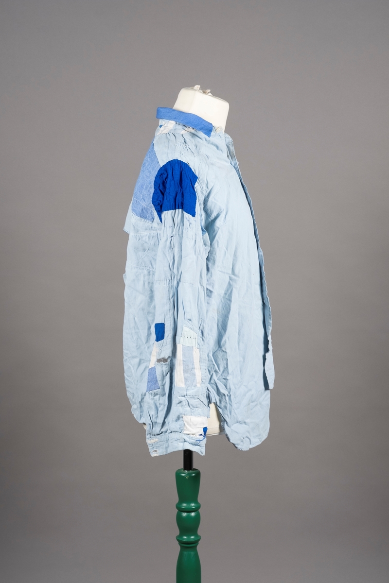 En lyselå skjorte reparert med påsydde lapper i andre blåfarger. Skjorten har tre knapper i plast, mangler to knapper. Det er brystlomme på høyre side og knapp på venstre mansjett. Fangenummer er påført under kragen på ryggen. Det kan være brodert en liten fisk over brystlommen.