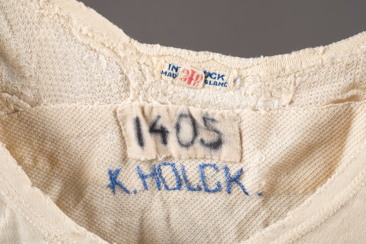 Hvit ullsinglet stoppet og lappet etter skader. Det er brodert inn initialer og navn på ulltrøyen i blå tråd. Noen av lappene har blomstermønstret stoff. Fangenummer er skrevet på i nakken.