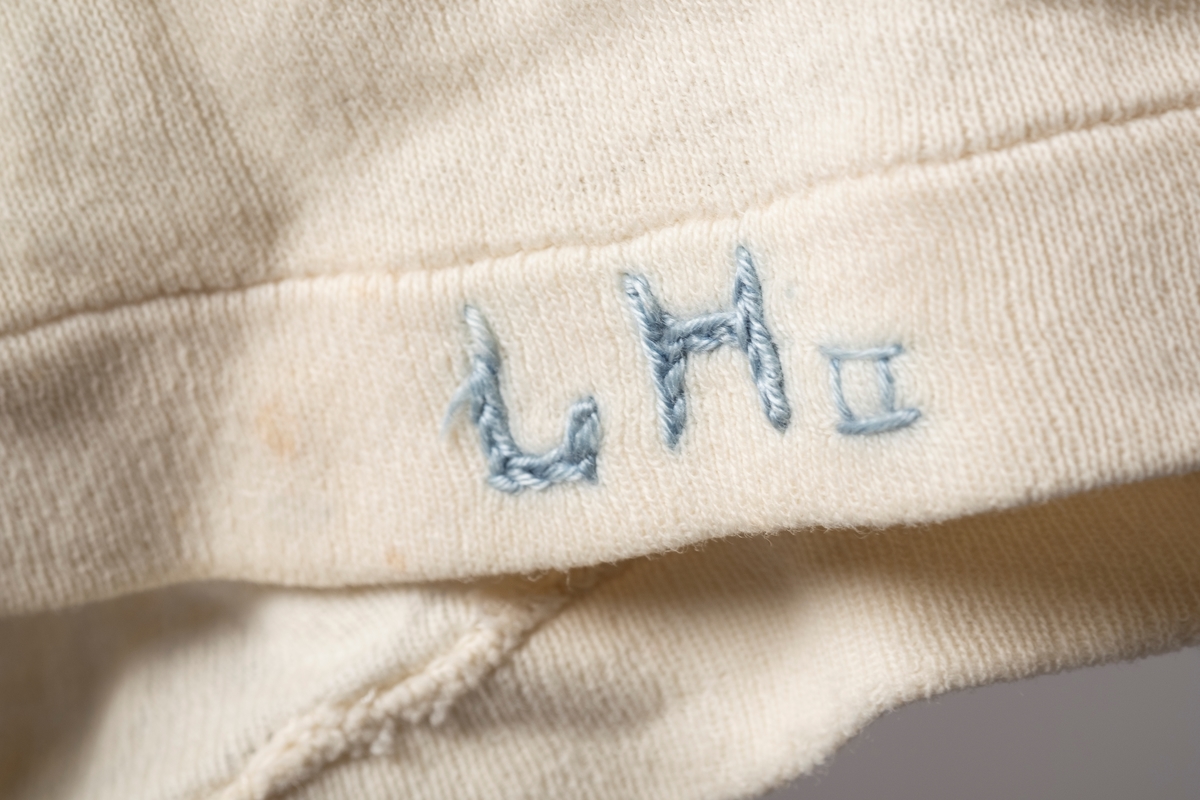 Hvit ullsinglet stoppet og lappet etter skader. Det er brodert inn initialer og navn på ulltrøyen i blå tråd. Noen av lappene har blomstermønstret stoff. Fangenummer er skrevet på i nakken.
