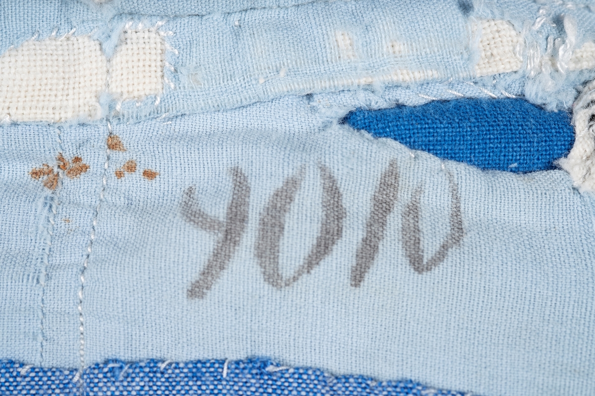 En lyselå skjorte reparert med påsydde lapper i andre blåfarger. Skjorten har tre knapper i plast, mangler to knapper. Det er brystlomme på høyre side og knapp på venstre mansjett. Fangenummer er påført under kragen på ryggen. Det kan være brodert en liten fisk over brystlommen.