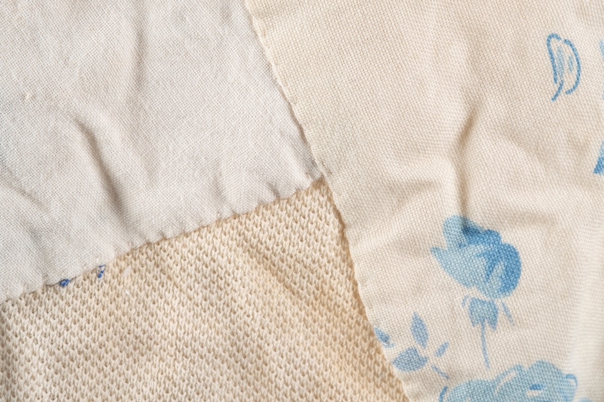 Hvit ullsinglet stoppet og lappet etter skader. Det er brodert inn initialer og navn på ulltrøyen i blå tråd. Noen av lappene har blomstermønstret stoff. Fangenummer er skrevet på i nakken.