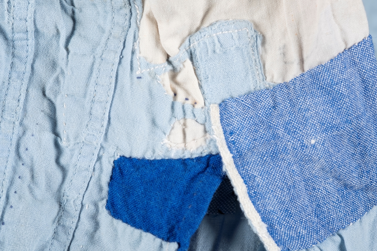 En lyselå skjorte reparert med påsydde lapper i andre blåfarger. Skjorten har tre knapper i plast, mangler to knapper. Det er brystlomme på høyre side og knapp på venstre mansjett. Fangenummer er påført under kragen på ryggen. Det kan være brodert en liten fisk over brystlommen.