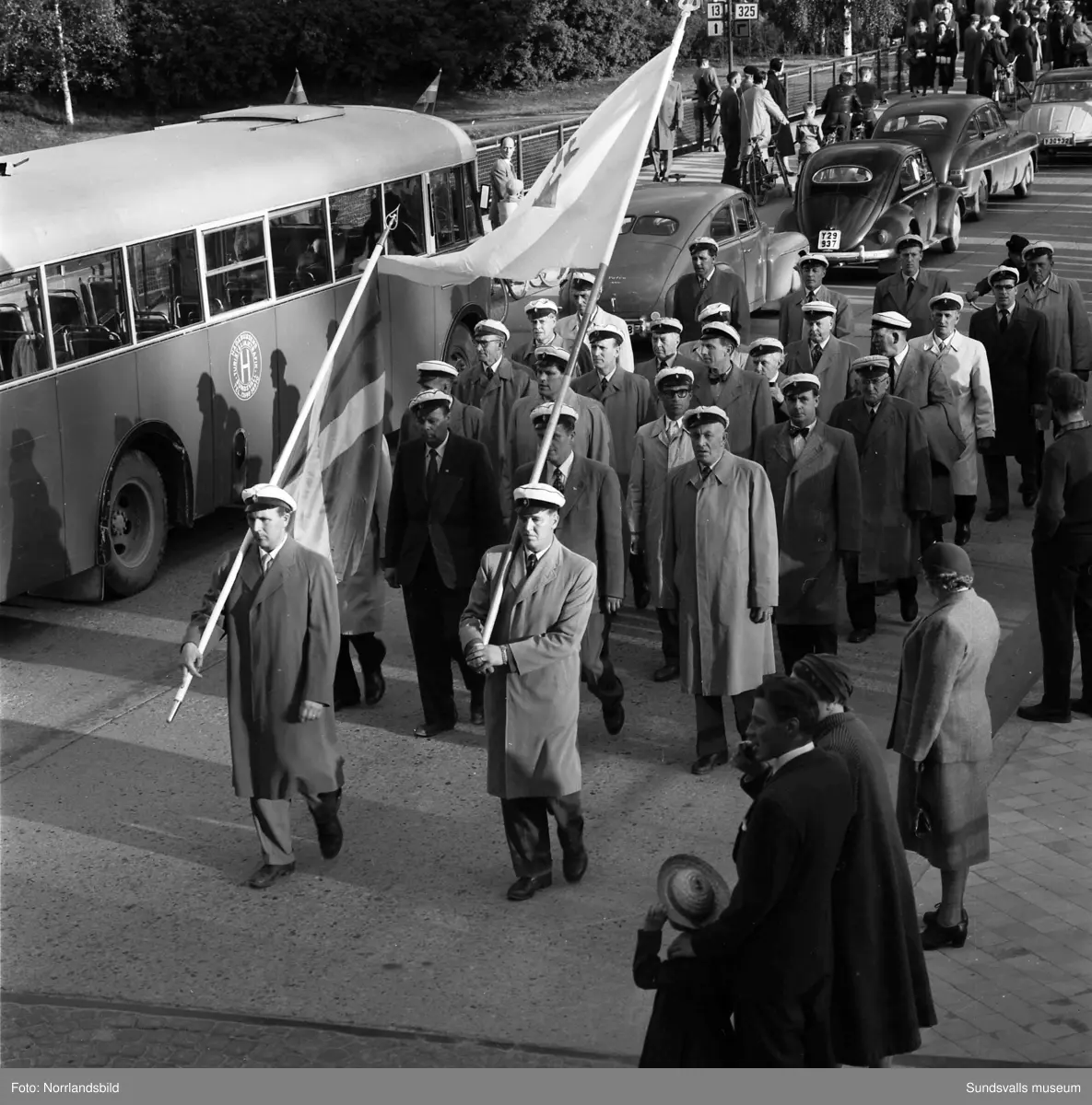 En stor serie bilder från firandet av Svenska flaggans dag 1956. Parad på Storbron med paradorkester, militär, scouter och mängder av olika föreningar och förbund. Därefter högtidlig uppställning i Idrottsparken med bland annat tal, körsång, gymnastikuppvisning och folkmusik.