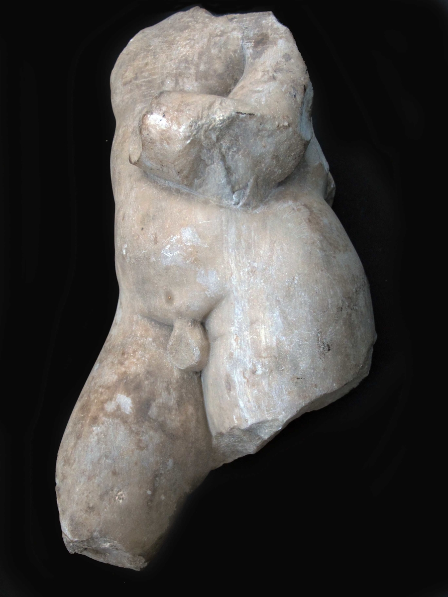 Torso av buespennende Eros, romersk keisertids kopi av gresk original.  Fin krystalinsk marmor. Figuren fragmentarisk bevart; hodet, høyre arm, vifigene og  bena delvis avslått. Figuren bøyer venstre arm opp mot brystet,  armen brukket av like over håndleddet. På bruddflaten et dypt boret hull, etter nagle som har holdt hånden fast. Figuren står på det venstre benet,  det høyre er skjøvet frem, og antageligvis har  kneet vært bøyet. Figuren knekker i hoften, og  dette gir den en s-formet bevegelse som går gjennom kroppen. Skulpturen er en romersk keisertidskopi av  den greske billedhugger Lysippos verk.