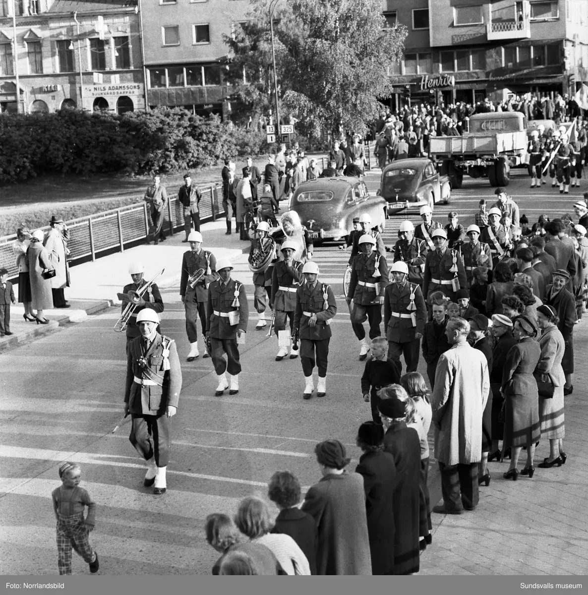 En stor serie bilder från firandet av Svenska flaggans dag 1956. Parad på Storbron med paradorkester, militär, scouter och mängder av olika föreningar och förbund. Därefter högtidlig uppställning i Idrottsparken med bland annat tal, körsång, gymnastikuppvisning och folkmusik.