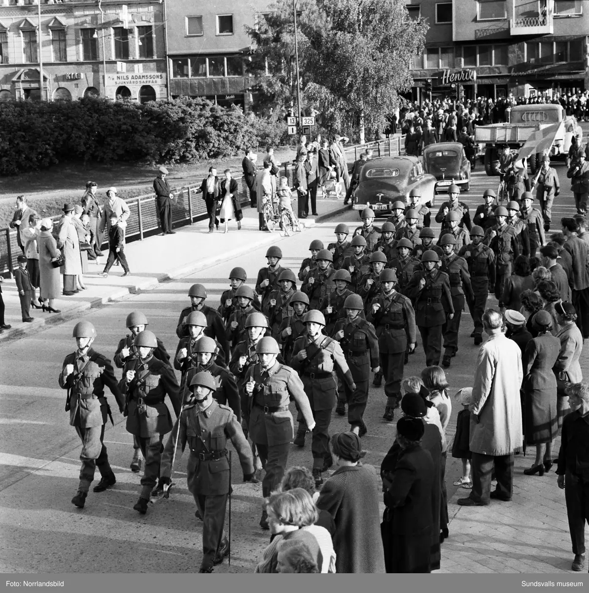 En stor serie bilder från firandet av Svenska flaggans dag 1956. Parad på Storbron med paradorkester, militär, scouter och mängder av olika föreningar och förbund. Därefter högtidlig uppställning i Idrottsparken med bland annat tal, körsång, gymnastikuppvisning och folkmusik.