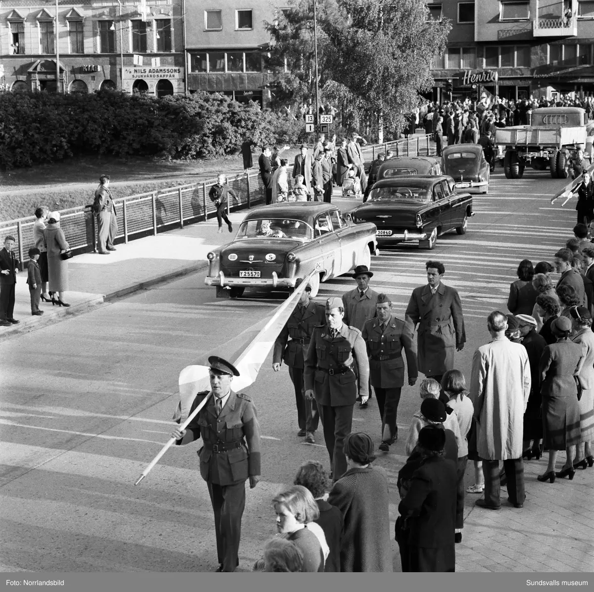 En stor serie bilder från firandet av Svenska flaggans dag 1956. Parad på Storbron med paradorkester, militär, scouter och mängder av olika föreningar och förbund. Därefter högtidlig uppställning i Idrottsparken med bland annat tal, körsång, gymnastikuppvisning och folkmusik.