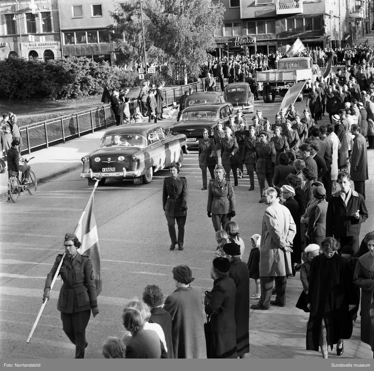 En stor serie bilder från firandet av Svenska flaggans dag 1956. Parad på Storbron med paradorkester, militär, scouter och mängder av olika föreningar och förbund. Därefter högtidlig uppställning i Idrottsparken med bland annat tal, körsång, gymnastikuppvisning och folkmusik.