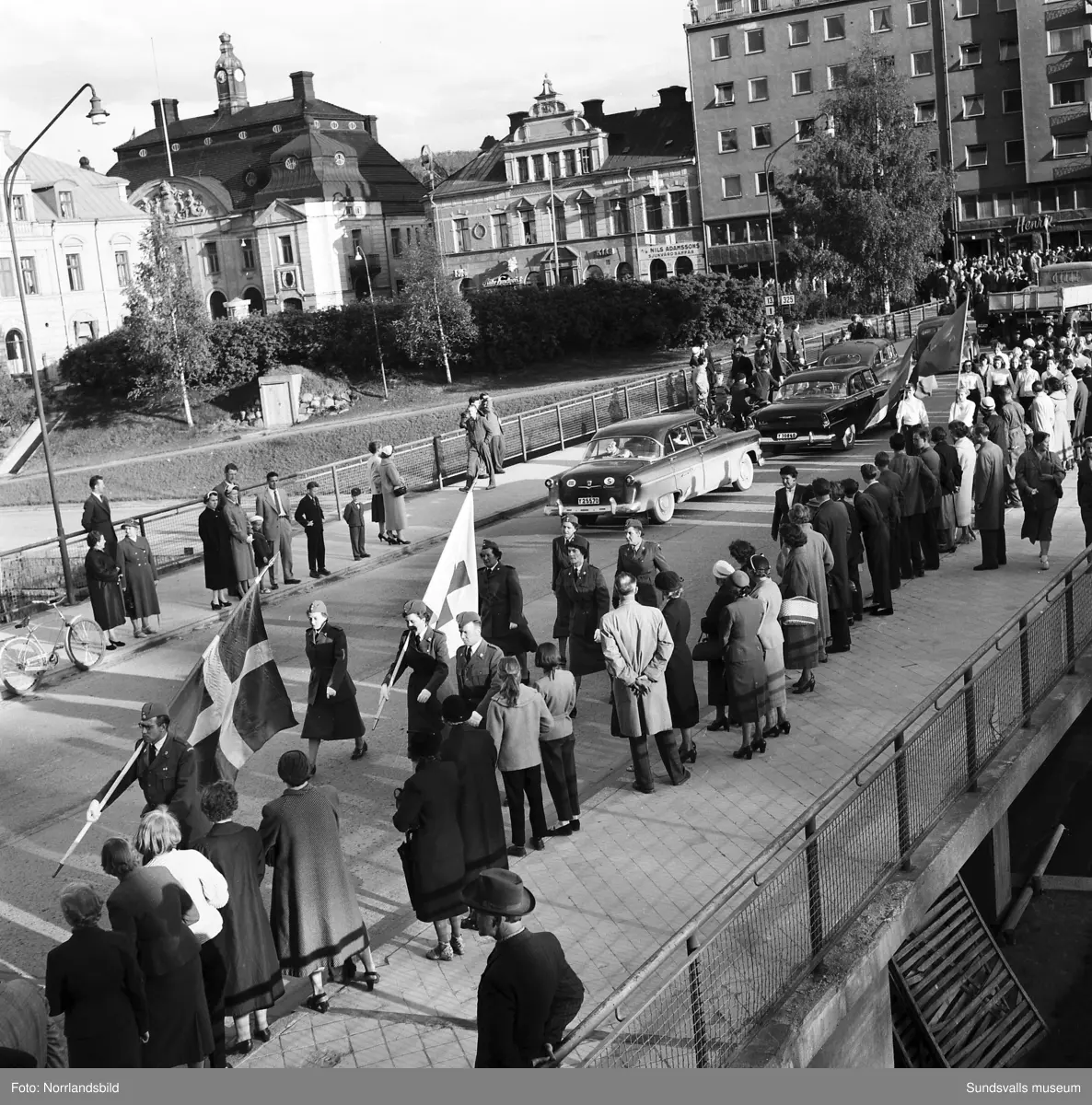 En stor serie bilder från firandet av Svenska flaggans dag 1956. Parad på Storbron med paradorkester, militär, scouter och mängder av olika föreningar och förbund. Därefter högtidlig uppställning i Idrottsparken med bland annat tal, körsång, gymnastikuppvisning och folkmusik.