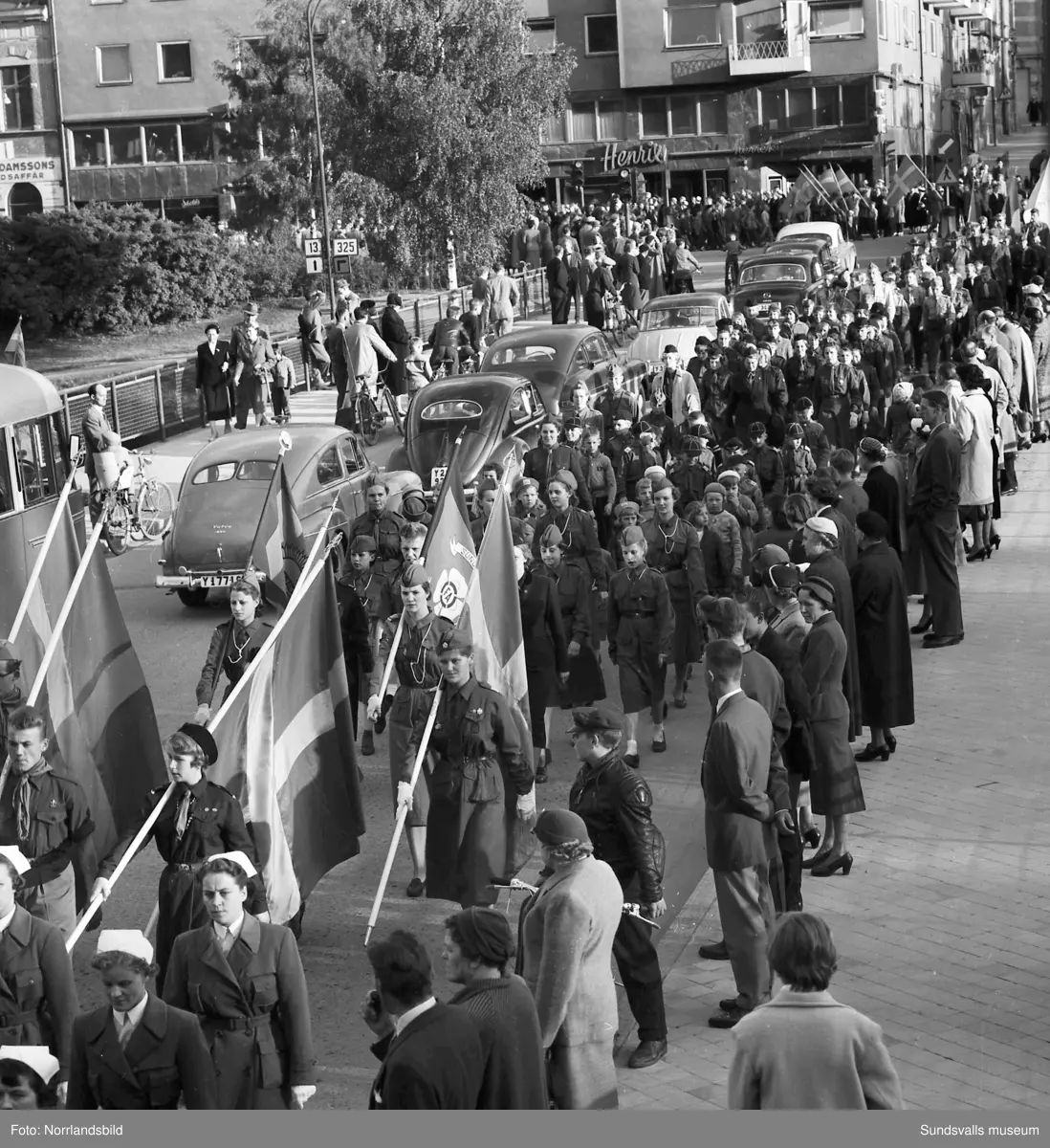 En stor serie bilder från firandet av Svenska flaggans dag 1956. Parad på Storbron med paradorkester, militär, scouter och mängder av olika föreningar och förbund. Därefter högtidlig uppställning i Idrottsparken med bland annat tal, körsång, gymnastikuppvisning och folkmusik.