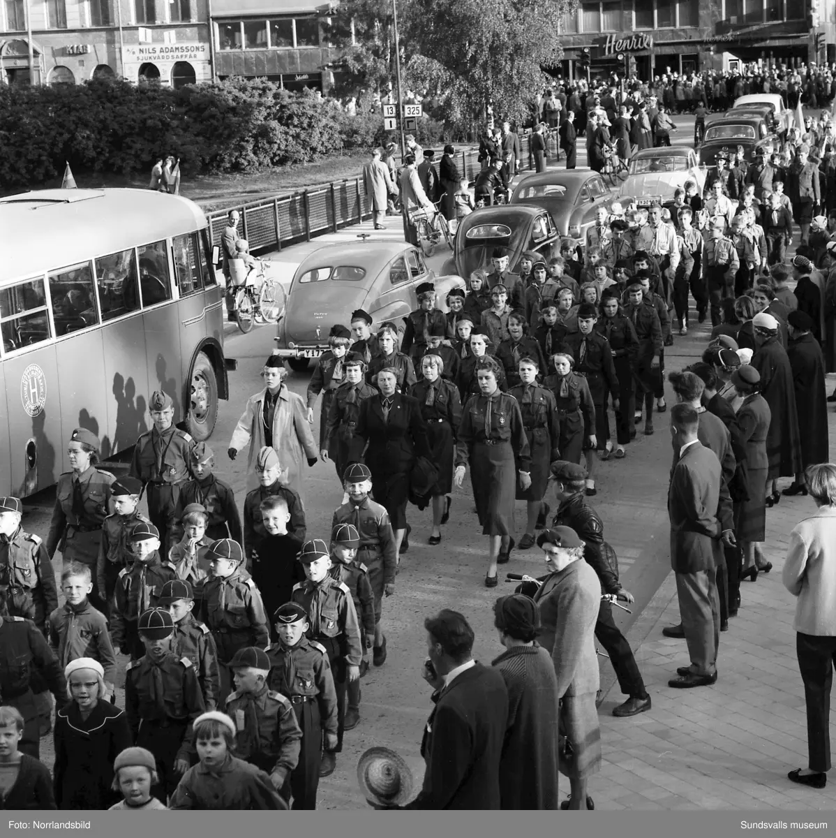 En stor serie bilder från firandet av Svenska flaggans dag 1956. Parad på Storbron med paradorkester, militär, scouter och mängder av olika föreningar och förbund. Därefter högtidlig uppställning i Idrottsparken med bland annat tal, körsång, gymnastikuppvisning och folkmusik.